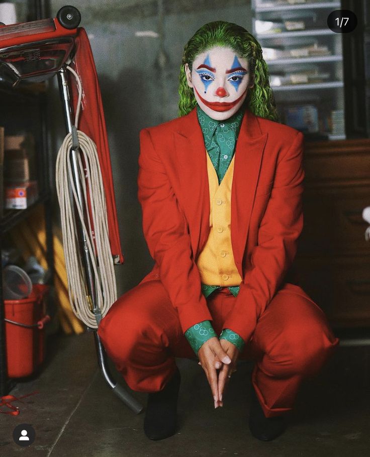 a man in a red suit and clown makeup sitting on the floor next to a shelf