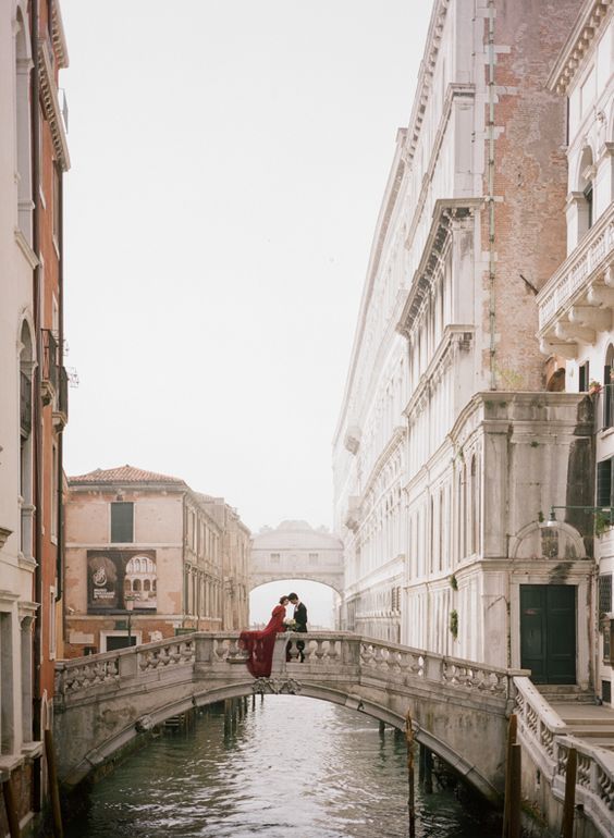 two people standing on a bridge over a canal in venice, italy with buildings lining both sides