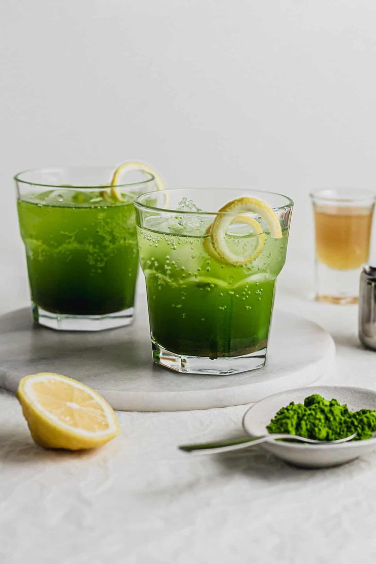 two glasses filled with green liquid next to lemon slices and herbs on a white surface