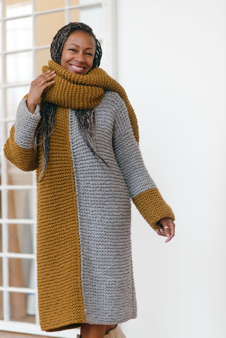 a smiling woman wearing a knitted scarf and coat, standing in front of a window