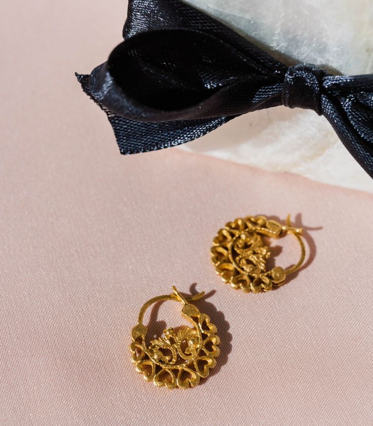 a pair of gold earrings sitting on top of a pink cloth next to a black bow