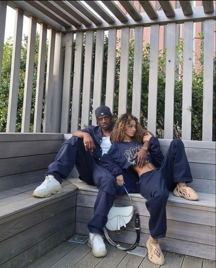 two people sitting on a wooden bench with their arms around each other and one person wearing a baseball cap