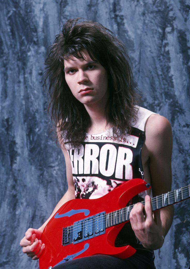 a woman with long hair holding a red guitar in front of a gray wall and looking at the camera