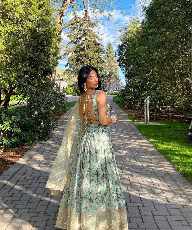 a woman in a long dress standing on a brick walkway with trees and bushes behind her