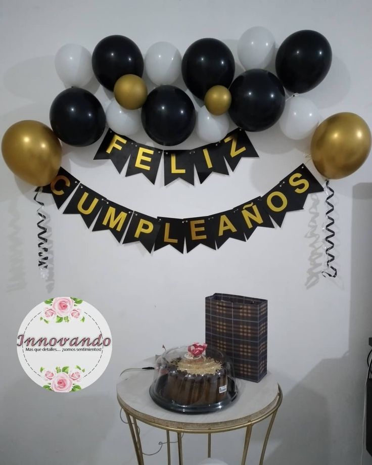 a table topped with a cake covered in black and gold balloons next to a sign that says feliv cumpleanos