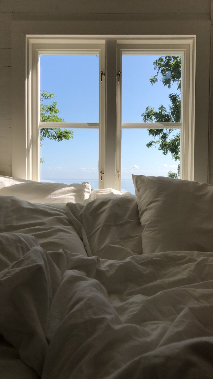 an unmade bed in front of two windows with white sheets and pillows on it