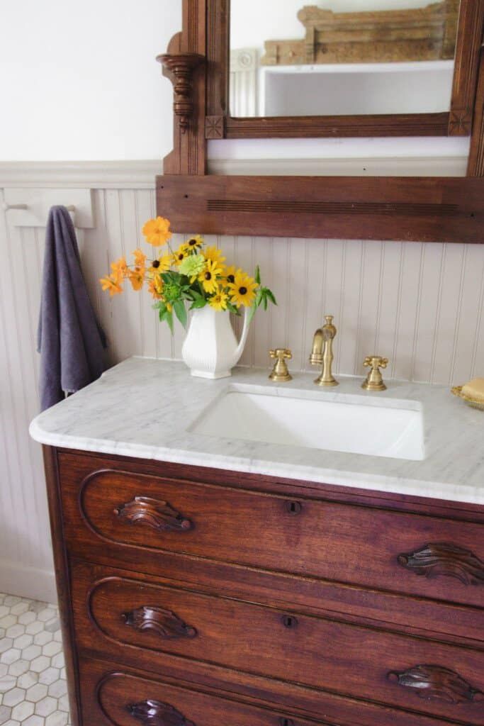 a white sink sitting under a mirror next to a wooden dresser with flowers in it