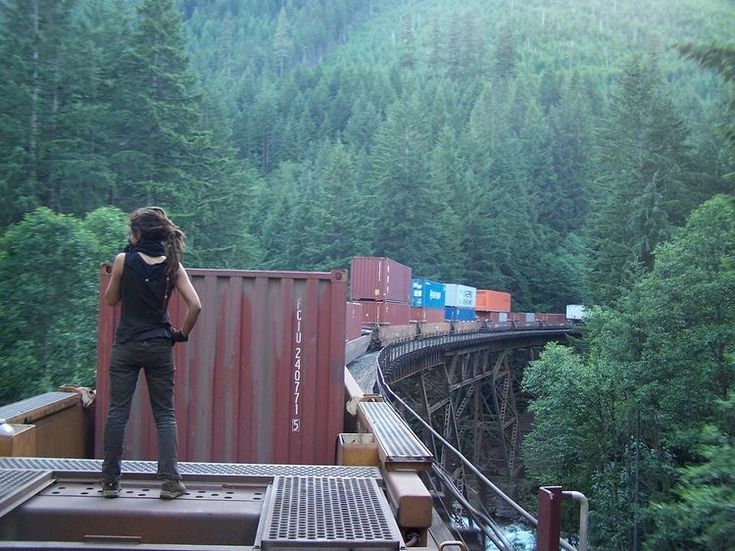 a woman standing on the side of a train track next to a lush green forest