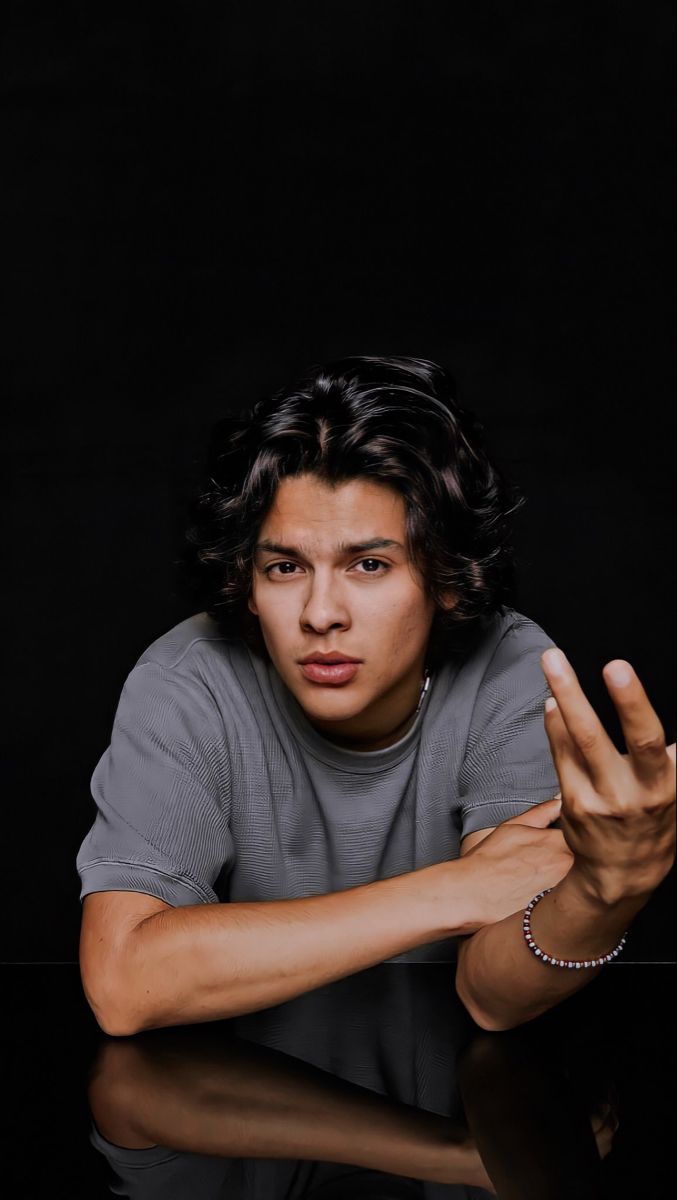 a young man sitting at a table in front of a black background holding his hands up