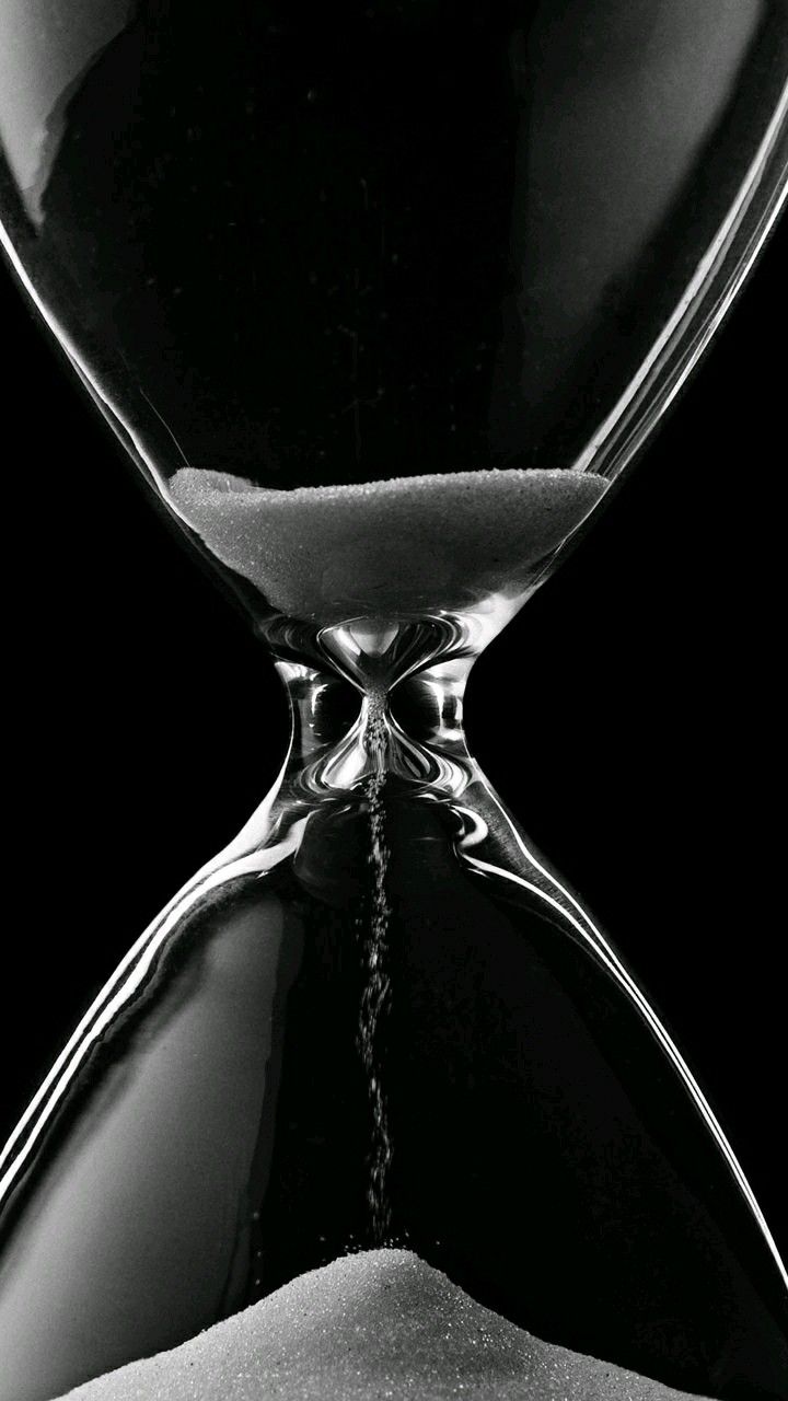 black and white photograph of an hourglass with sand running through the middle, in front of a dark background