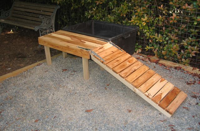 a wooden bench sitting next to a trash can on top of a gravel ground near a fence