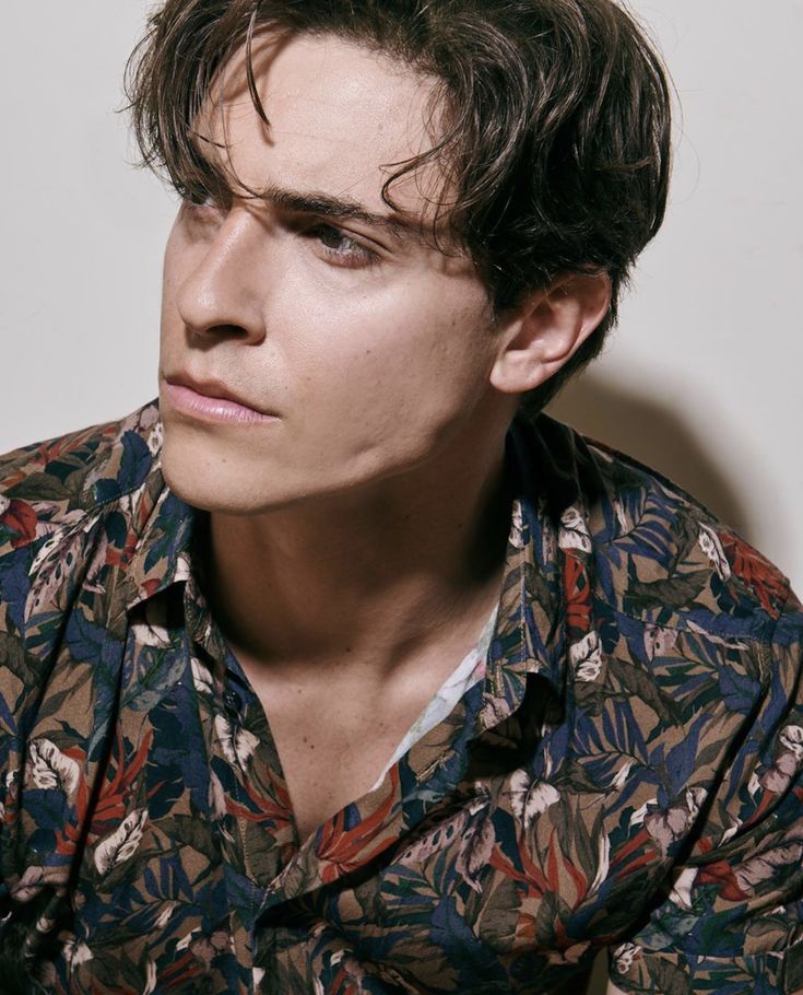 a man with dark hair wearing a floral shirt and looking off to the side while standing in front of a white wall