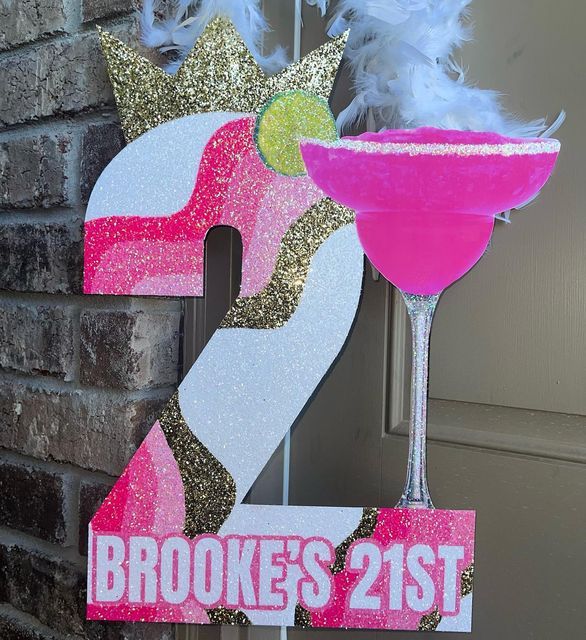 a pink and gold 21st birthday sign next to a brick wall with a drink in it