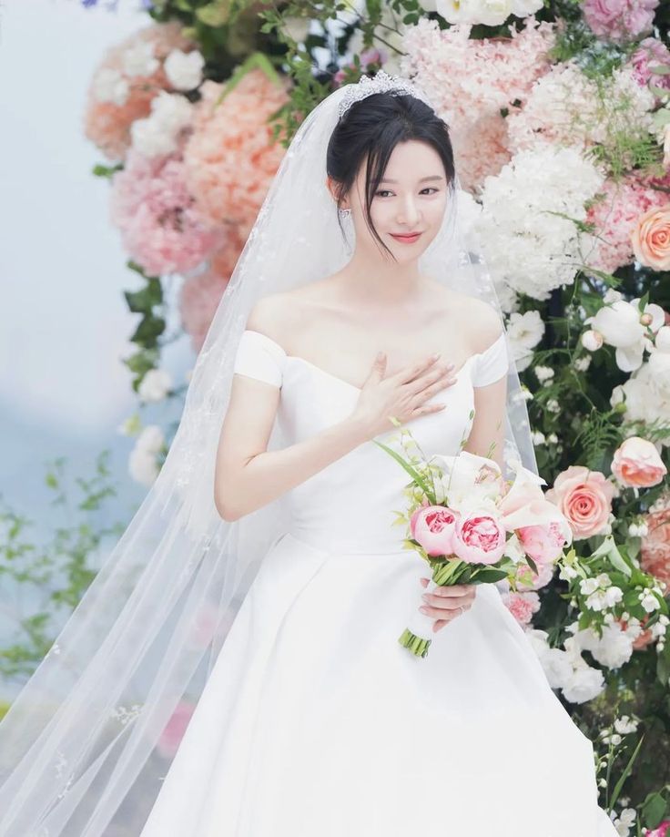 a woman in a white wedding dress standing next to a flower covered wall with pink and white flowers