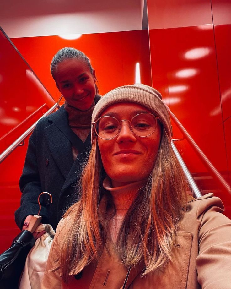 a woman wearing glasses and a hat taking a selfie in front of a red wall