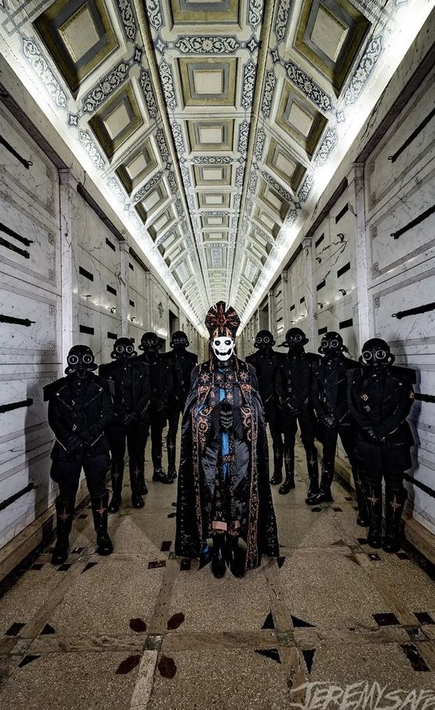 a group of masked people walking down a long hallway with white walls and ceiling tiles