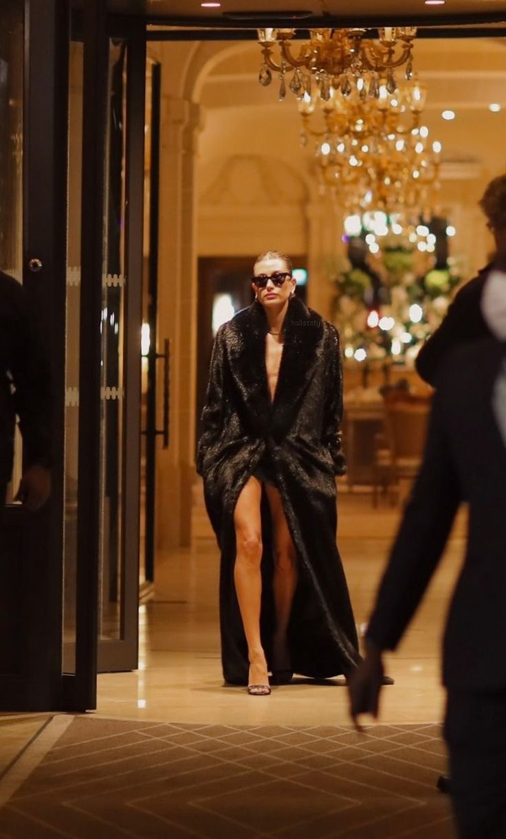 a woman in a black coat and sunglasses walking into a building with a chandelier hanging from the ceiling