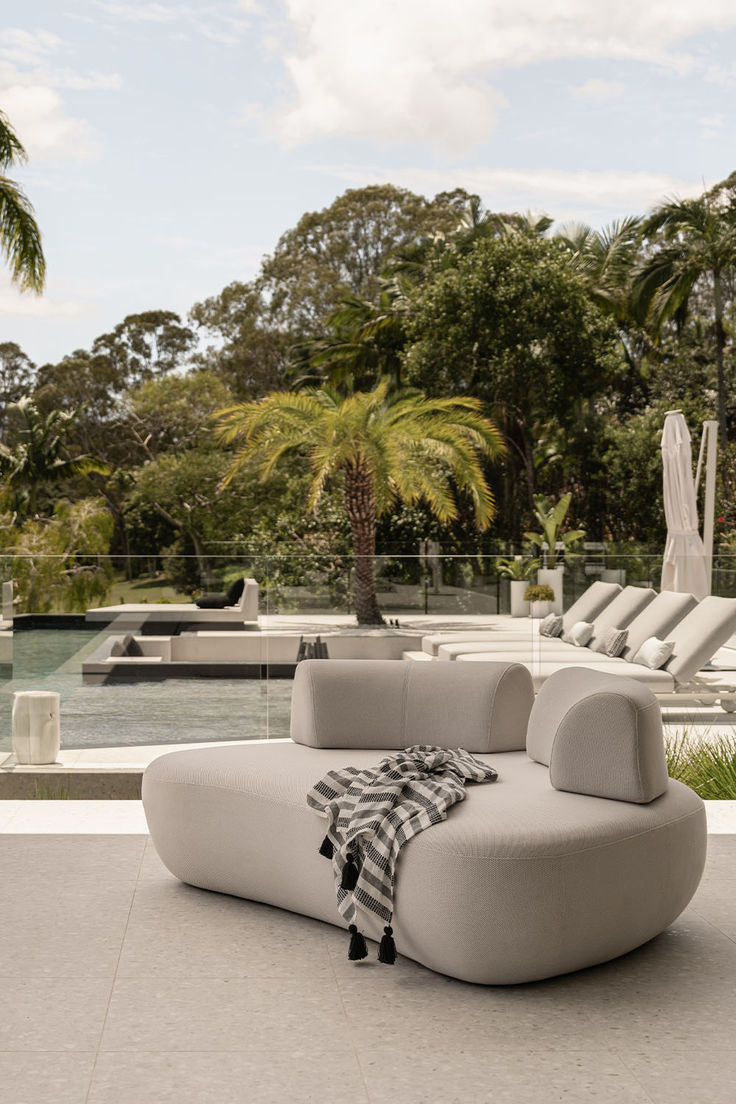 a white couch sitting on top of a cement floor next to a palm tree filled pool