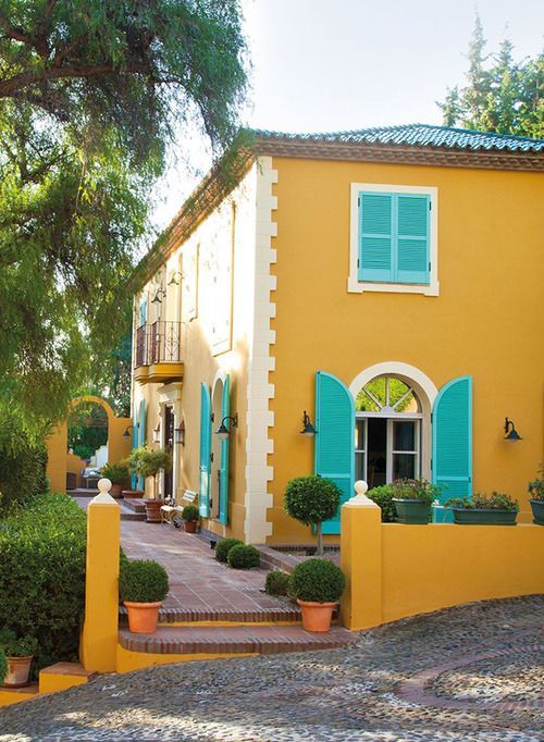 a yellow house with green shutters and blue windows on the outside, surrounded by greenery