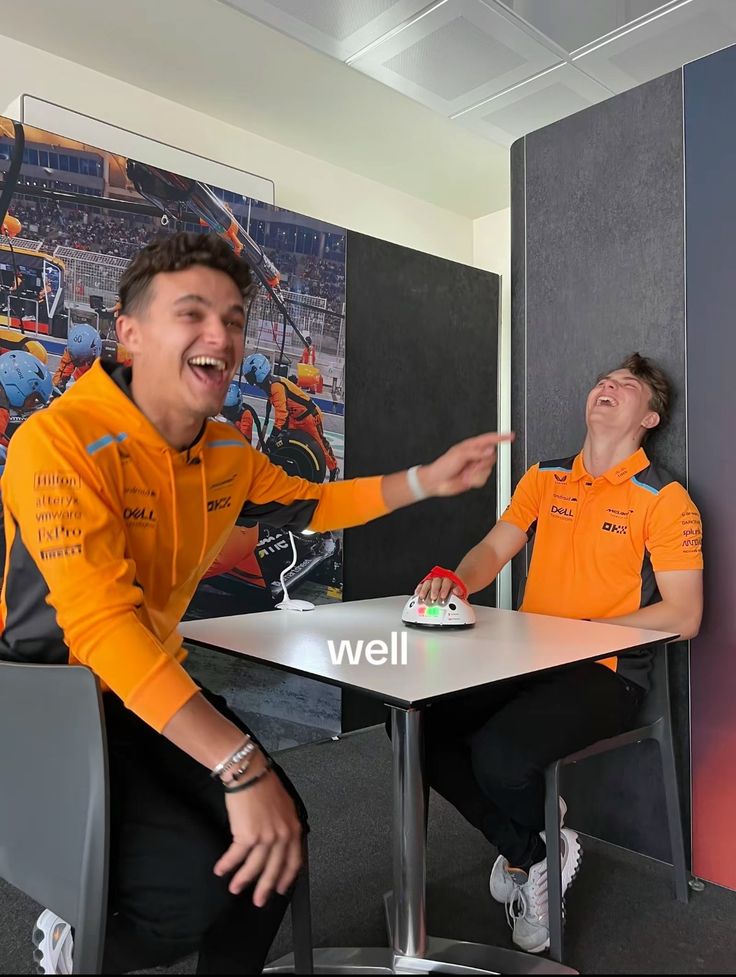 two men in orange shirts sitting at a table and pointing to each other with their hands