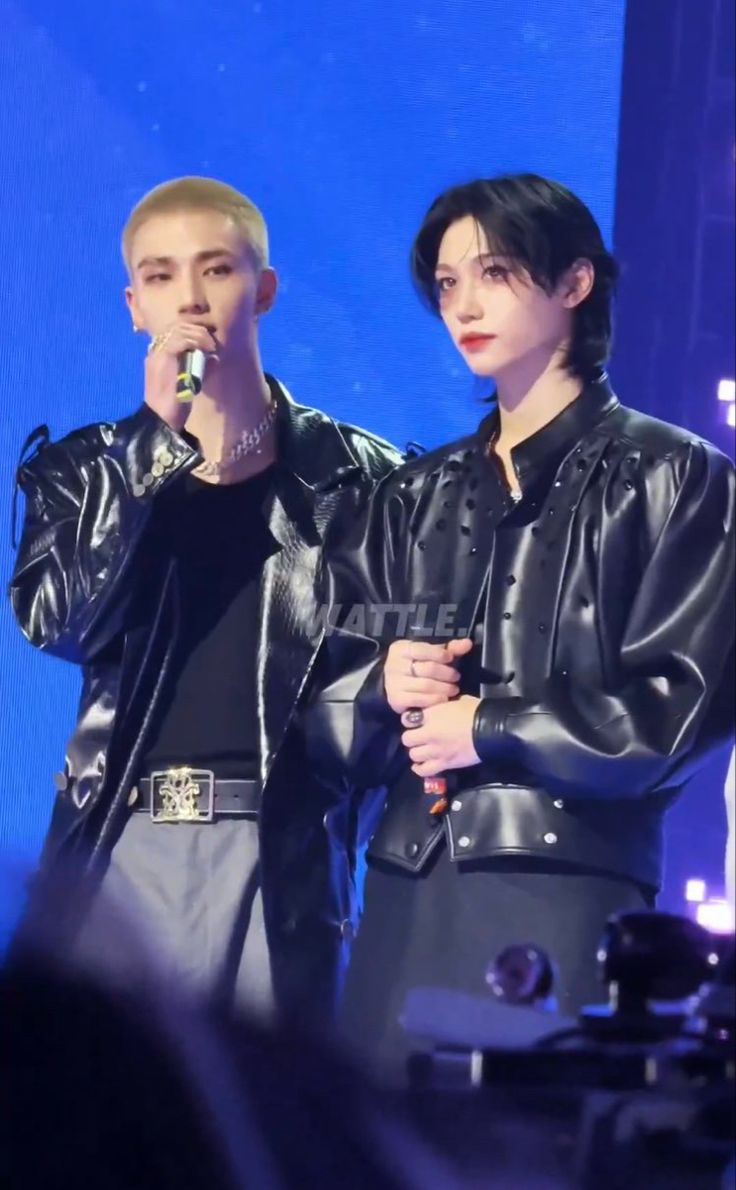 two people standing next to each other on stage with microphones in their hands and one person wearing a black leather jacket