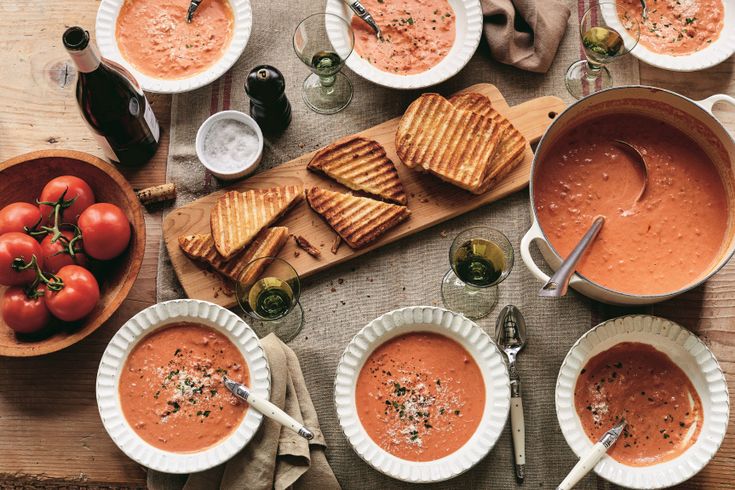 a table topped with bowls of soup next to grilled cheese and tomato slices on top of a cutting board