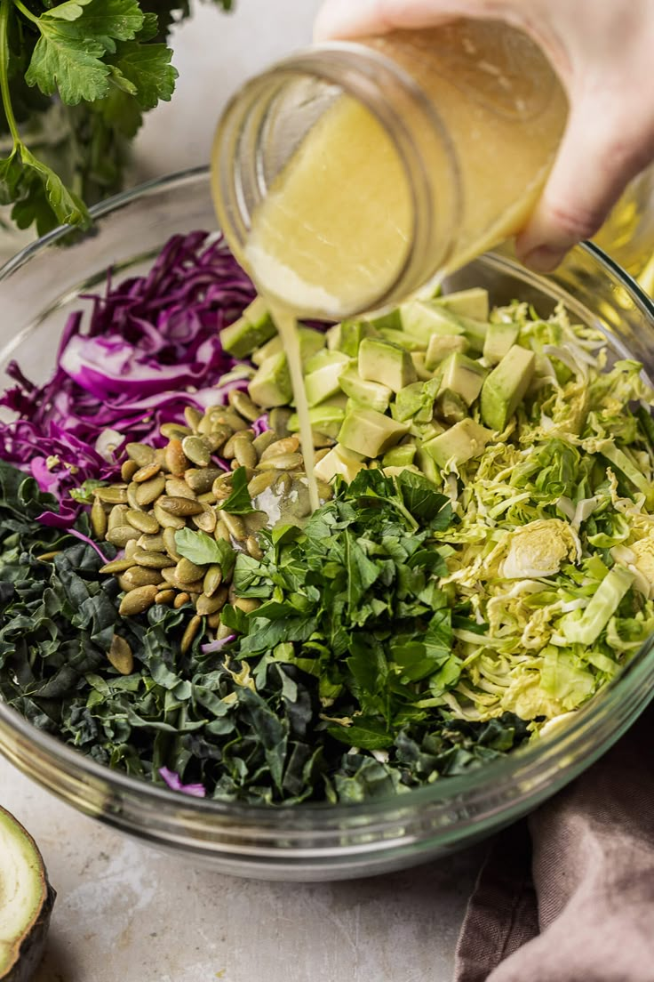 someone pouring dressing into a glass bowl filled with chopped vegetables and avocado slices