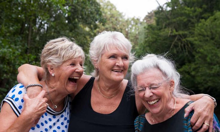 three older women standing next to each other in front of some trees and bushes with their arms around one another