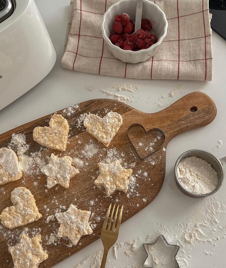 some heart shaped cookies are on a cutting board next to a bowl of raspberries