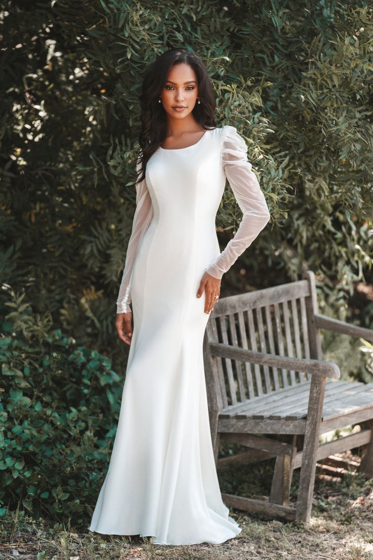 a woman standing in front of a bench wearing a long sleeved white dress with sheer sleeves