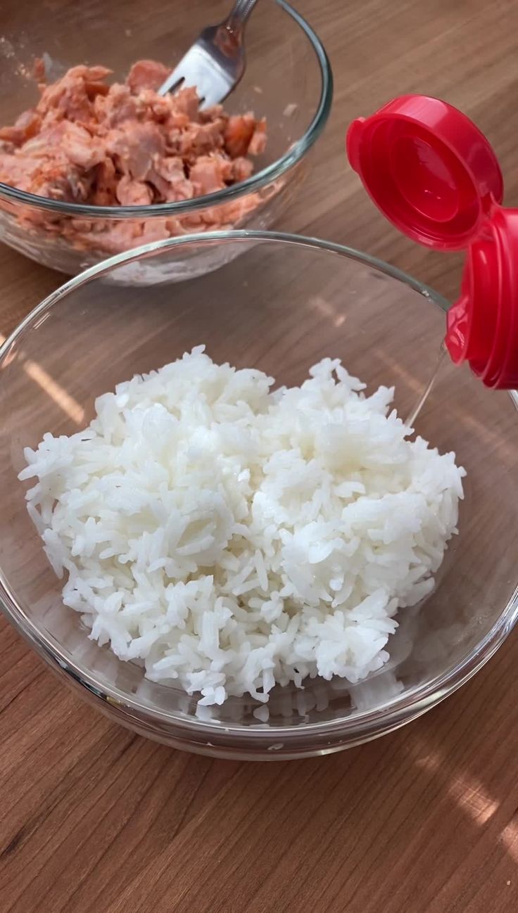 two bowls filled with rice and meat on top of a wooden table next to a red pepper shaker