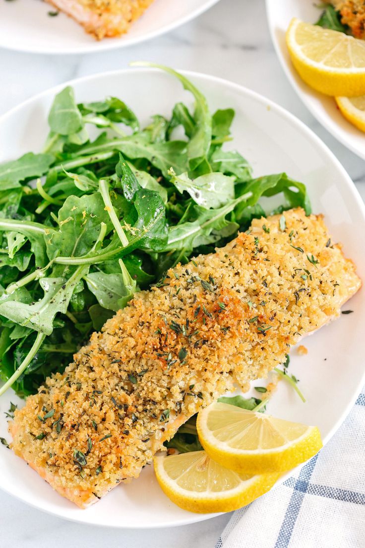 two plates filled with fish and greens on top of a white table cloth next to sliced lemons