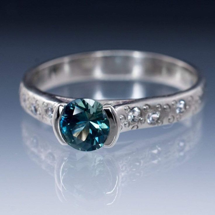 Teal to blue Fair Trade Montana sapphire engagement ring with a semi or half bezel setting. The ring shank will crusted with a star dust like texture of flush set white sapphires and tiny engraved circles. The fair trade genuine sapphire will be handset and the ring has a satin-brushed finish as pictured ( polished finish is available upon request). The band width is 3mm. Height above the finger about 6 mm. It's a Fair Trade Level 1 Gemstone, the highest level available. The ring pictures were t Stylish Engagement Rings, Montana Sapphire Engagement, Montana Sapphire Engagement Ring, Moissanite Engagement Ring White Gold, Half Bezel, Star Dust, Vintage Engagement Rings Unique, Oval Cut Engagement Ring, Teal Sapphire