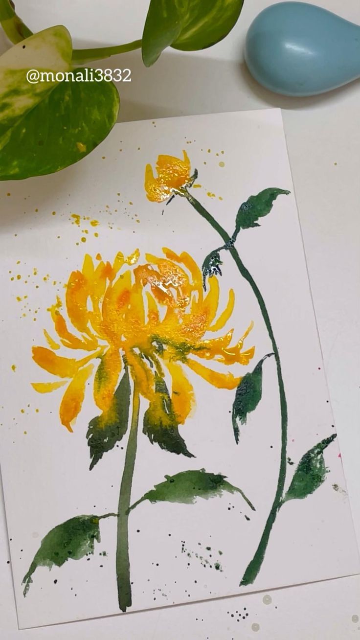 a yellow flower painted on white paper next to some green leaves and a blue spoon