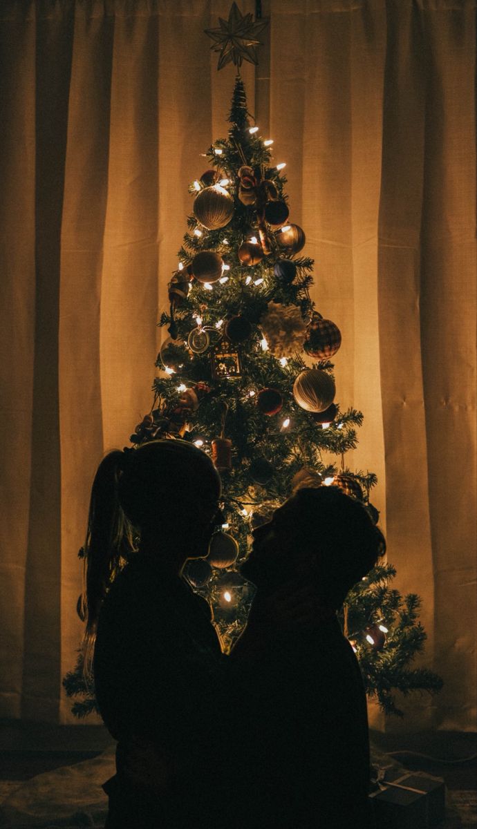 two people sitting in front of a christmas tree with lights on it and a dog standing next to them