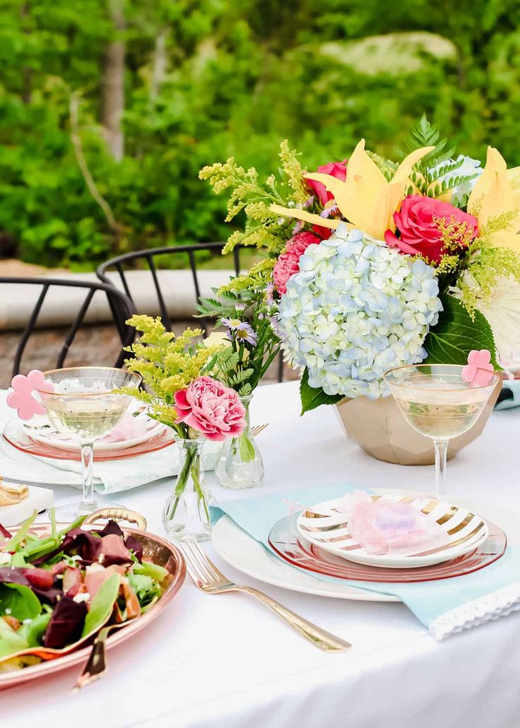 a table set with plates and flowers in vases on top of it, next to a bowl of salad