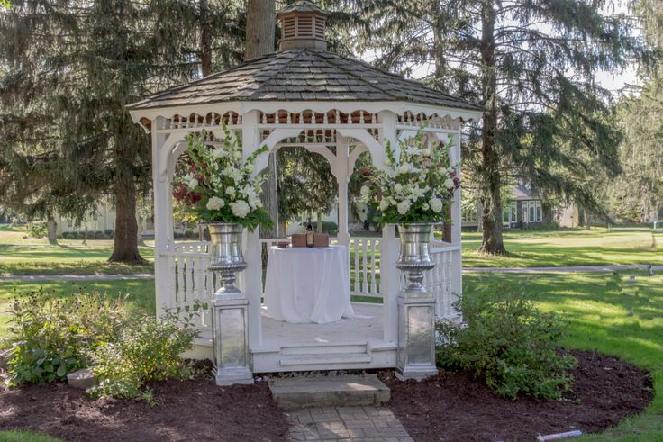 a white gazebo with flowers on it in the middle of some grass and trees