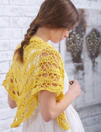 a woman standing in front of a brick wall wearing a yellow crochet shawl