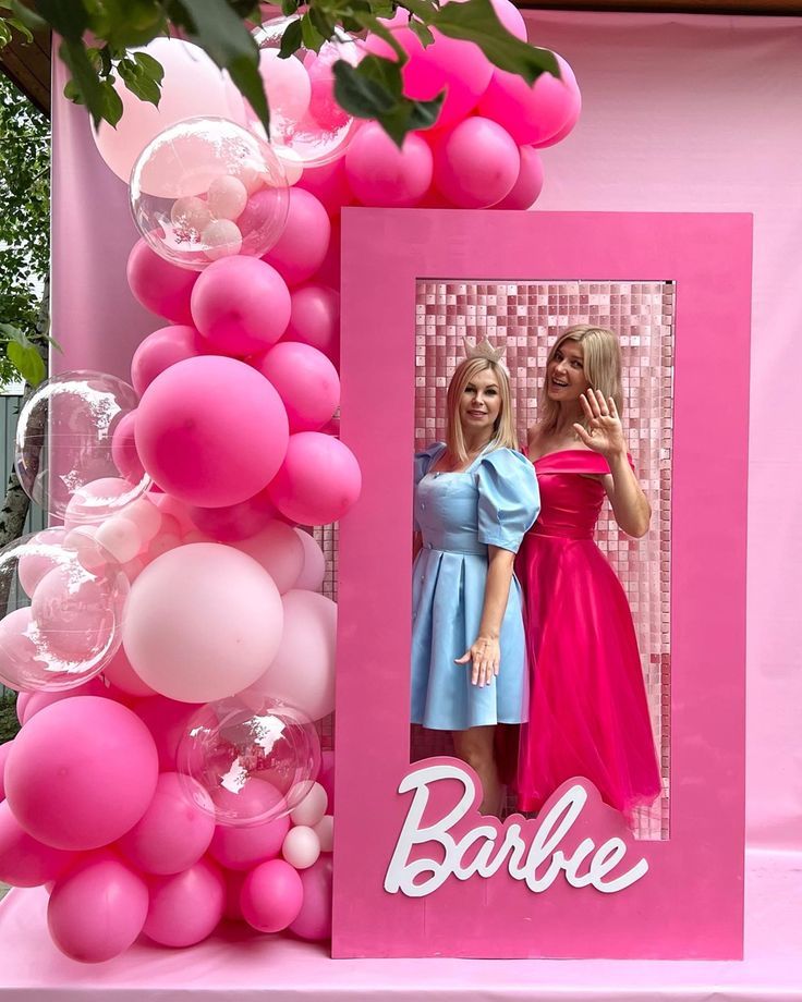 two women are standing in front of a pink photo frame with balloons hanging from it