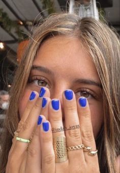 a woman covering her face with both hands and looking at the camera while wearing blue nail polish