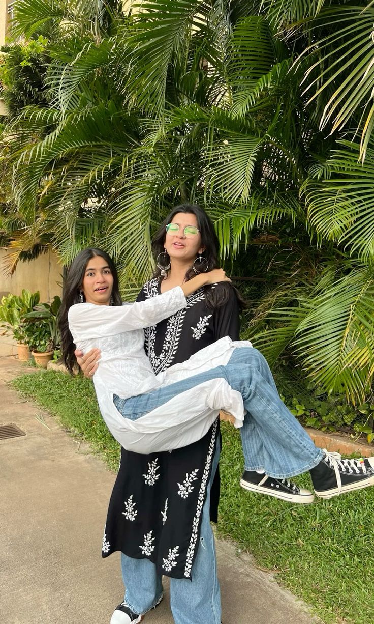 two women standing next to each other in front of palm trees and bushes, one carrying the woman on her back