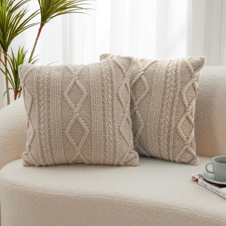 two knitted pillows sitting on a couch next to a coffee cup and book with a plant in the background