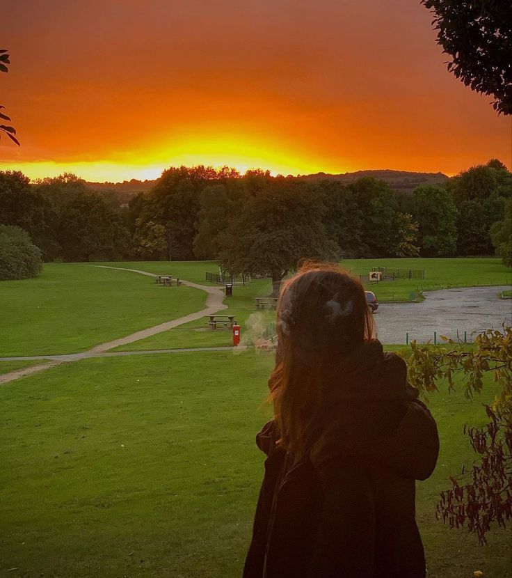 a woman standing on top of a lush green field next to a park at sunset