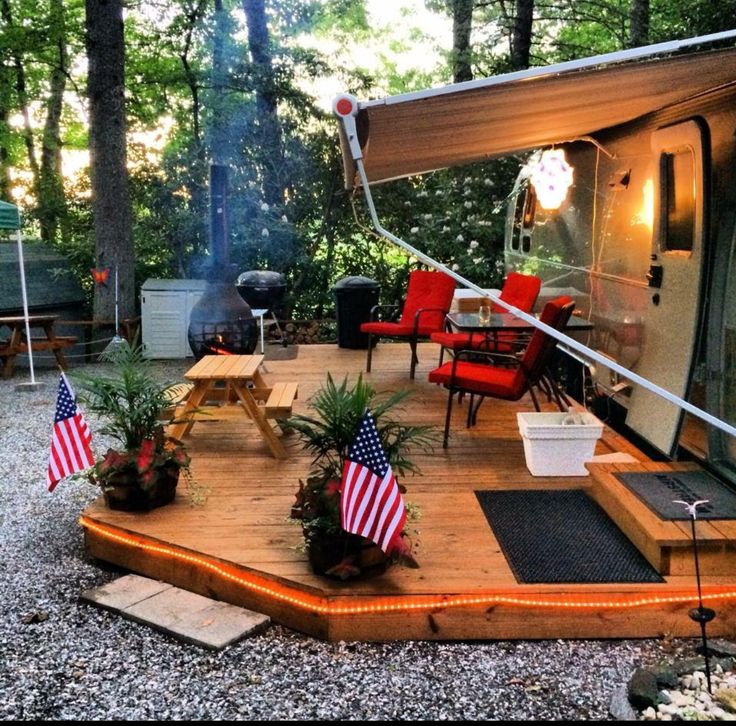 an outdoor deck with flags and lights on it, next to a camper trailer