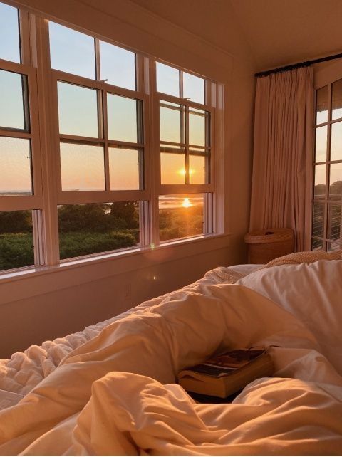 an unmade bed with a book on it in front of two large windows at sunset