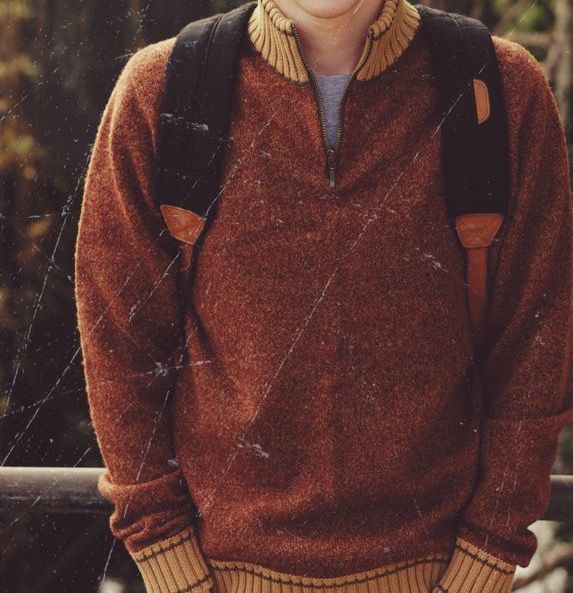 a young boy wearing a brown sweater and suspenders standing in front of a fence