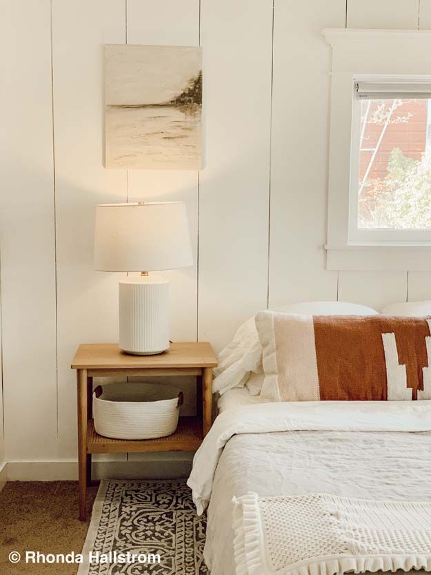 a white bed sitting under a window next to a night stand with a lamp on it