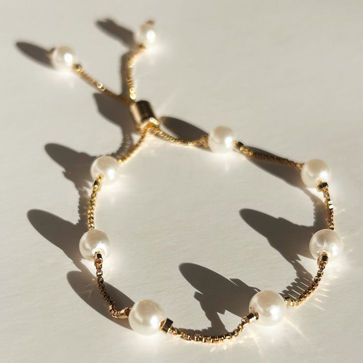 a bracelet with white pearls and gold chains on it's side, sitting on a table