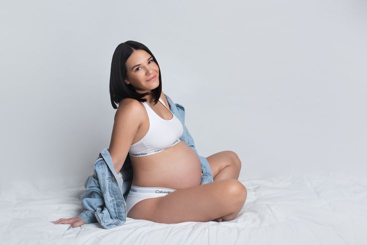 a pregnant woman sitting on top of a bed wearing a white bra and jean jacket