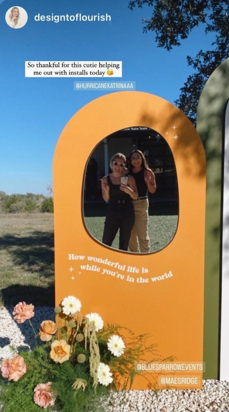 two women taking a selfie in front of an orange sign with flowers on it